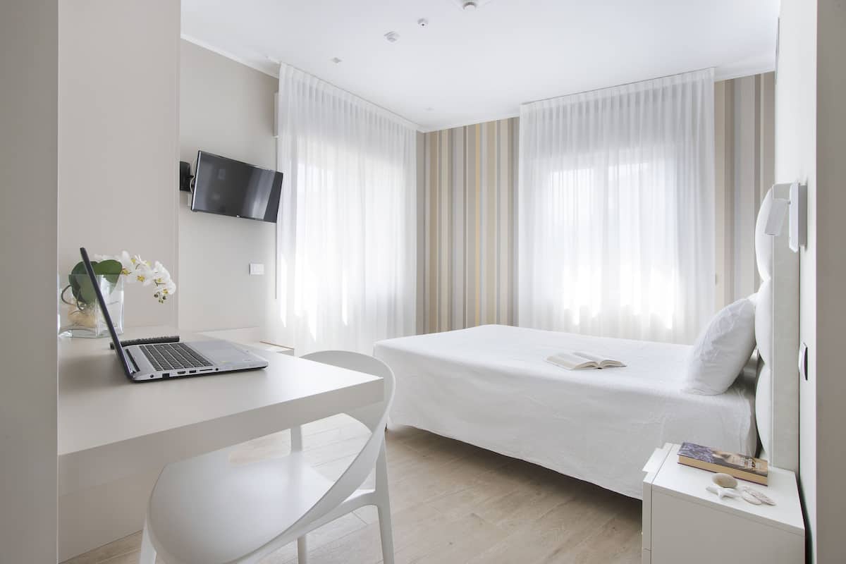 A modern hotel room with a bed, a desk with a laptop, a TV mounted on the wall, and a bedside table with a book and a remote control.