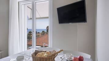Uma área de refeições com uma mesa posta para dois, uma cesta de pão, duas maçãs e uma televisão na parede.