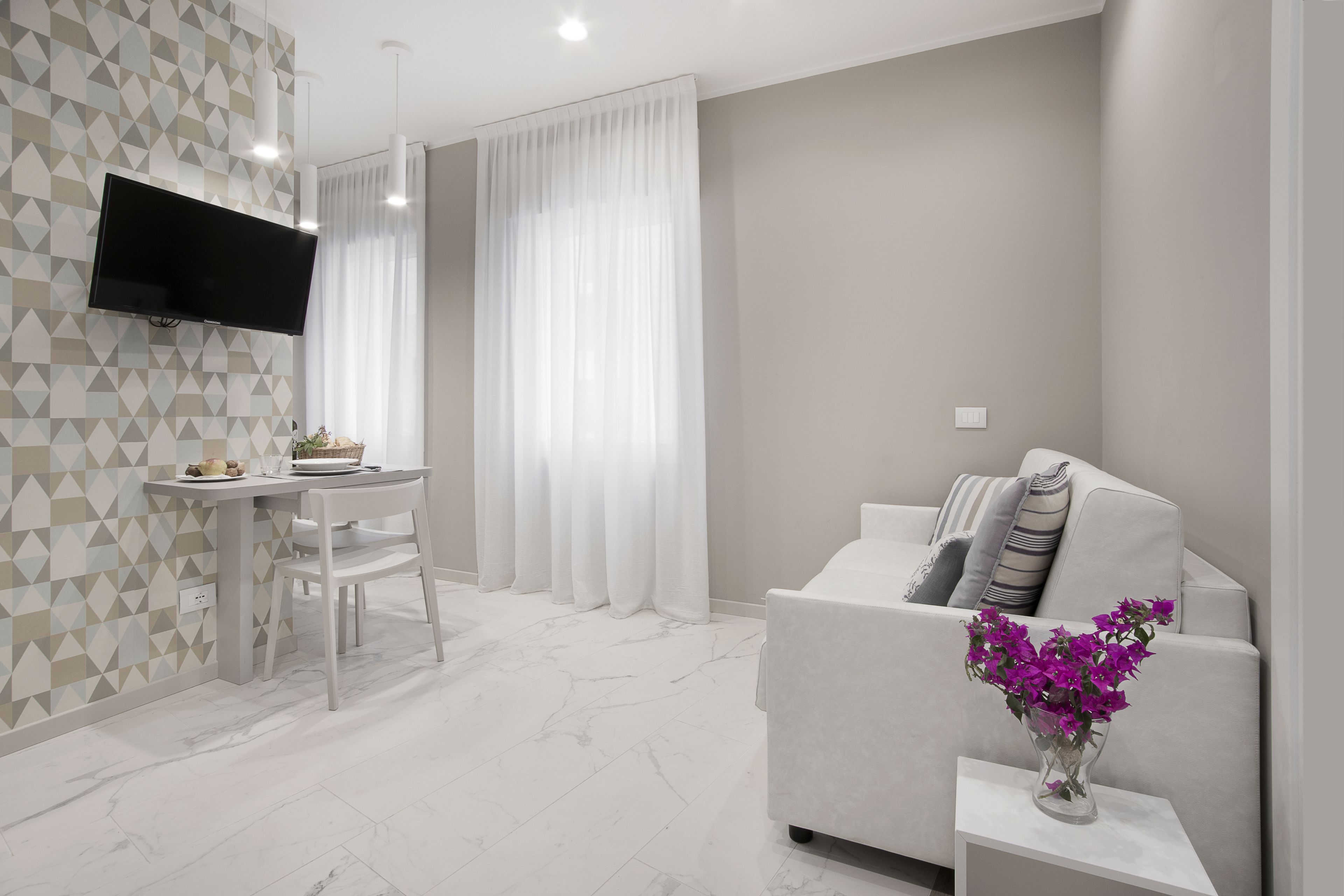 Un soggiorno moderno con una TV a schermo piatto montata a parete, un'area pranzo con tavolo e sedie, e un divano con un vaso di fiori viola.