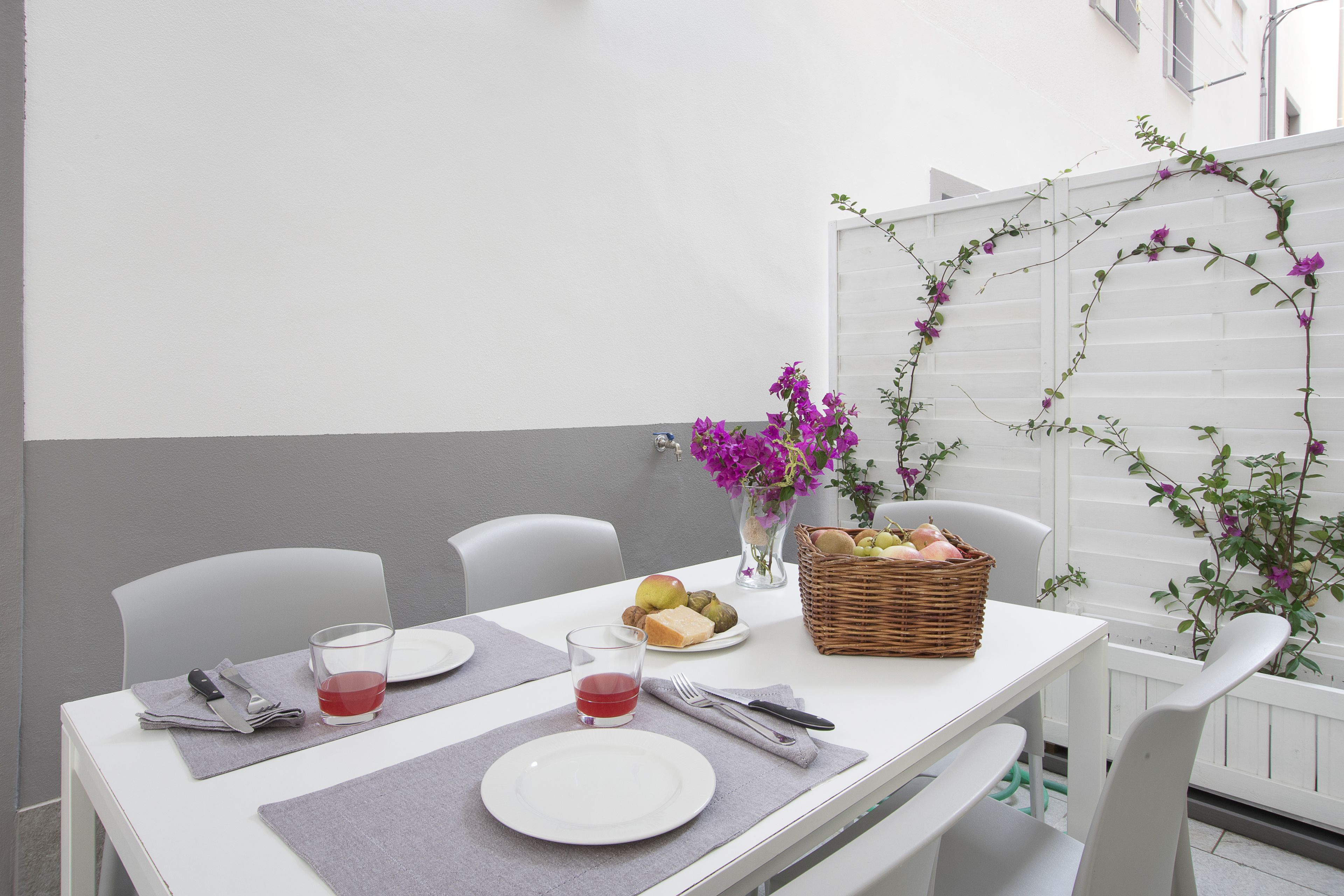 Una mesa dispuesta para dos, con platos, cubiertos y una copa de bebida roja. Hay una cesta de mimbre con frutas y un jarrón con flores moradas.