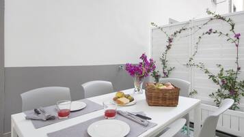 Una mesa dispuesta para dos, con platos, cubiertos y una copa de bebida roja. Hay una cesta de mimbre con frutas y un jarrón con flores moradas.