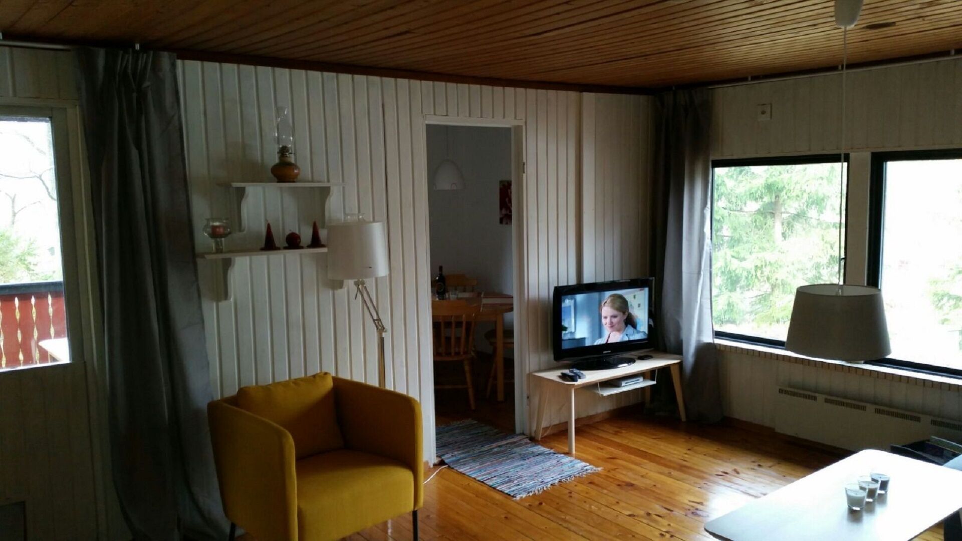 A living room with a television, a yellow armchair, a dining area, and large windows.