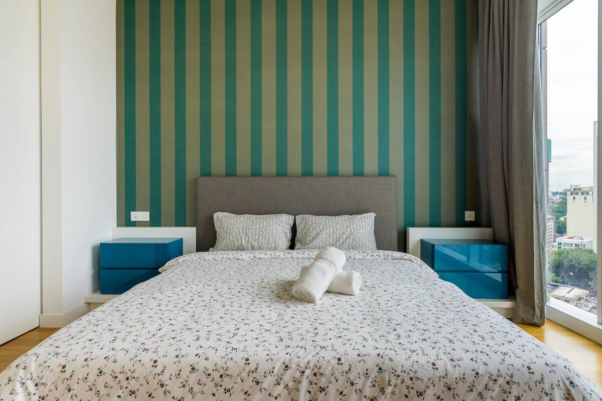 A neatly made bed with floral bedding, a striped wall, and a city view through a window.