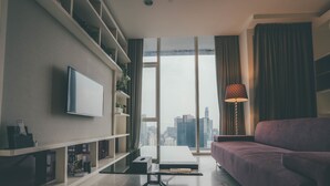 A modern living room with a flat-screen TV mounted on a white shelf, a purple sofa, and a large window offering a city view. - LUMA At Platinum Suite (Kuala Lumpur)