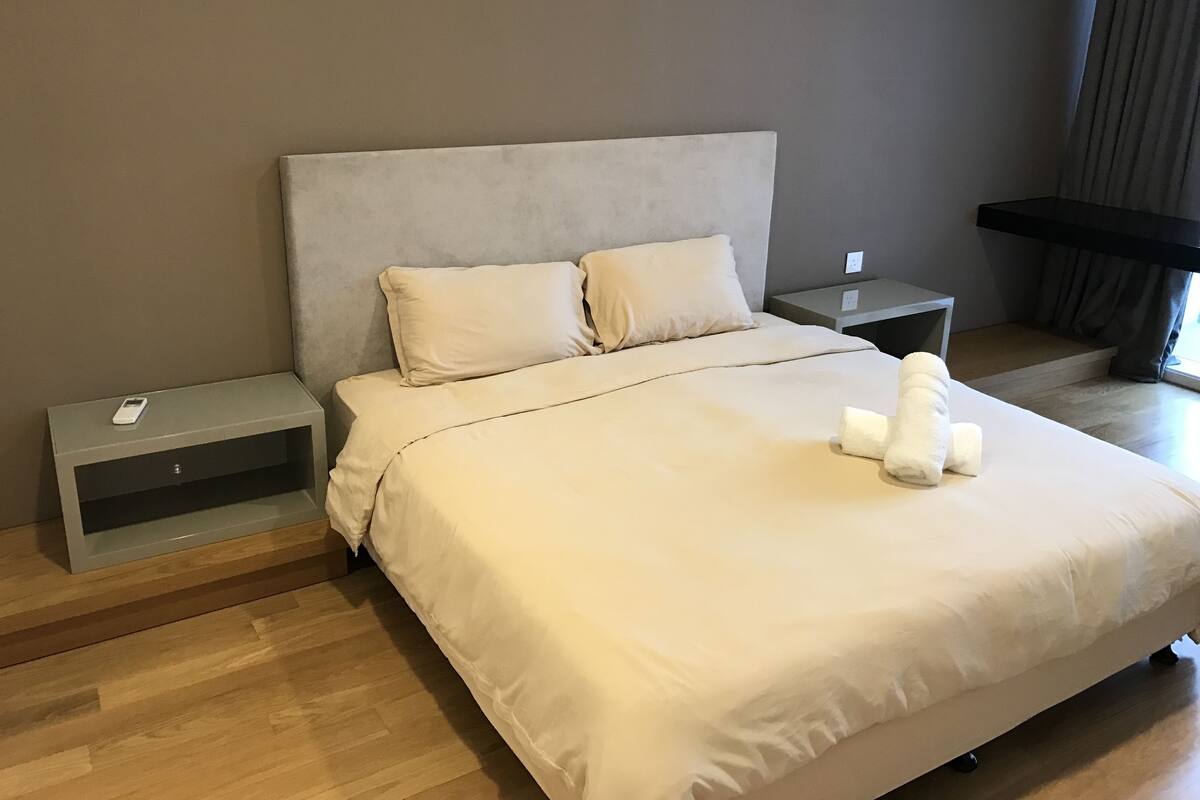 A neatly made bed with a white comforter and pillows, a bedside table with a lamp, and a window with curtains.