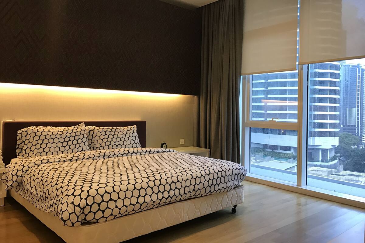 A modern bedroom with a large bed, a nightstand, and a view of tall buildings through a large window.
