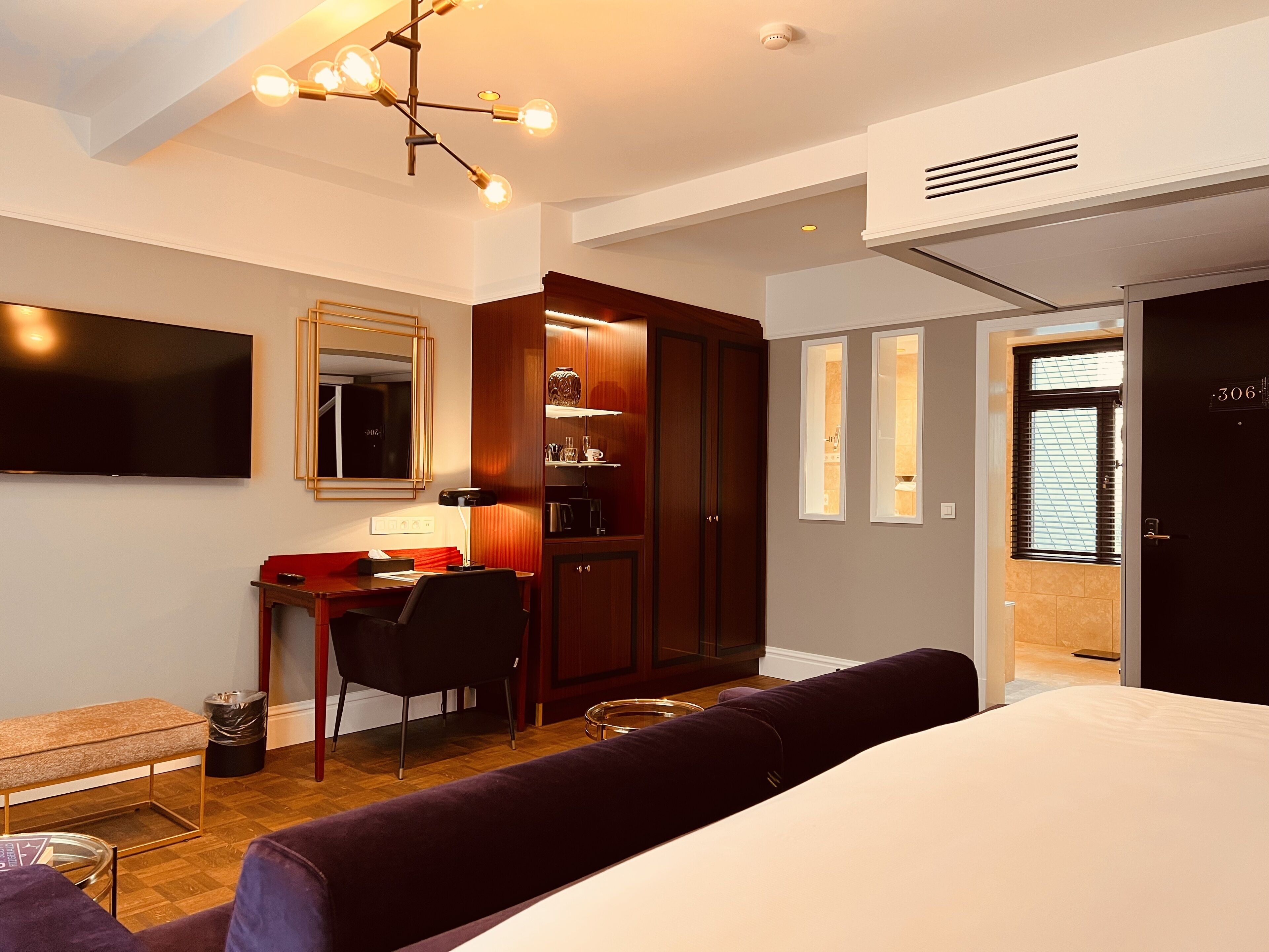 Una habitación de hotel moderna con una cama, un escritorio con una silla, un televisor, un armario y un baño visible a través de una puerta abierta.