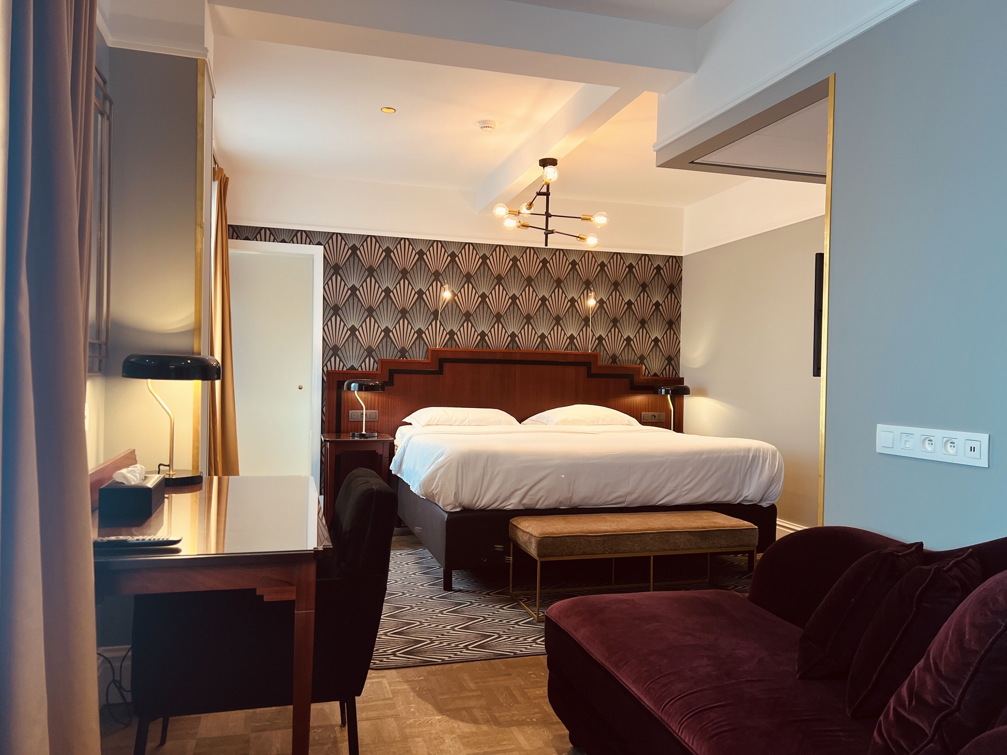 Habitación de hotel con cama, escritorio, silla, banco, pared con estampado y lámpara de techo.