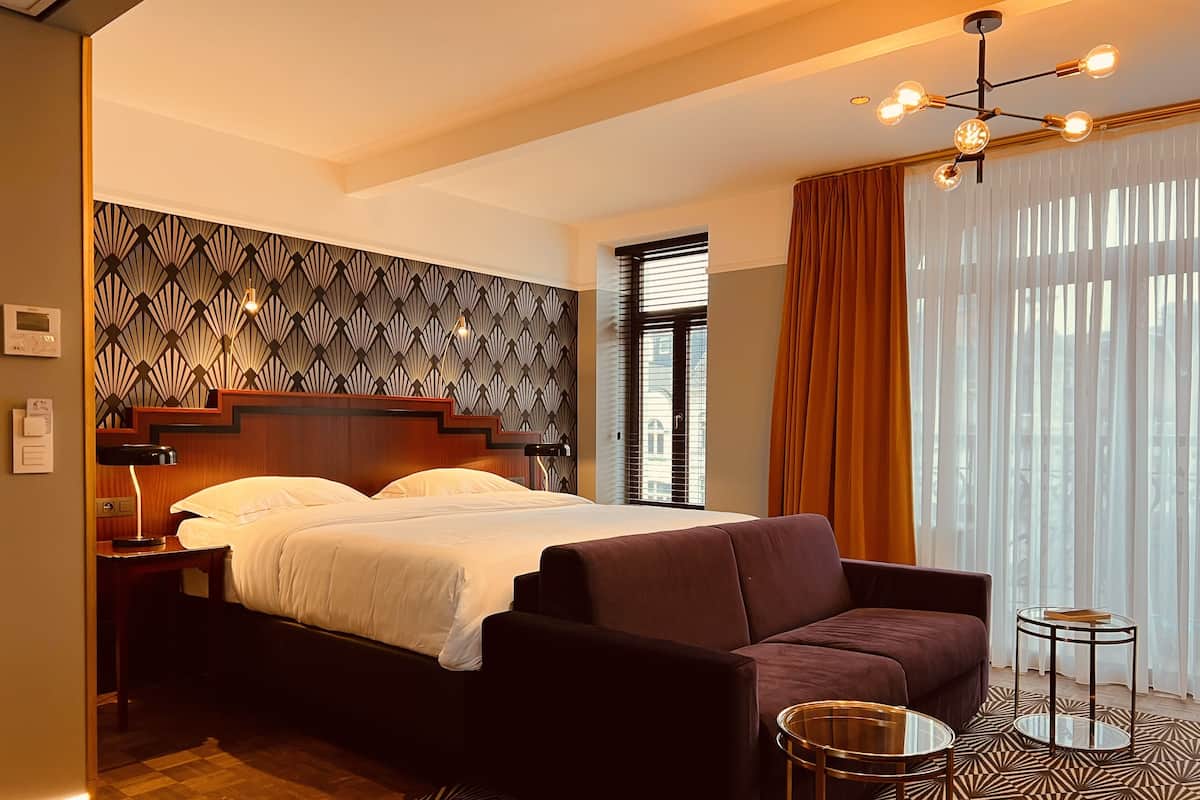 Une chambre d’hôtel moderne avec un grand lit, un fauteuil sombre, une petite table ronde et un tapis à motifs.