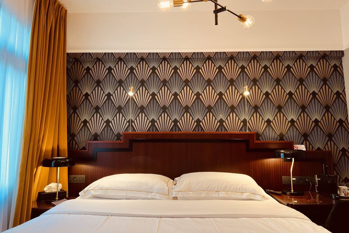 Une chambre d’hôtel avec un grand lit, un papier peint à motifs, un lustre moderne et une tête de lit en bois.