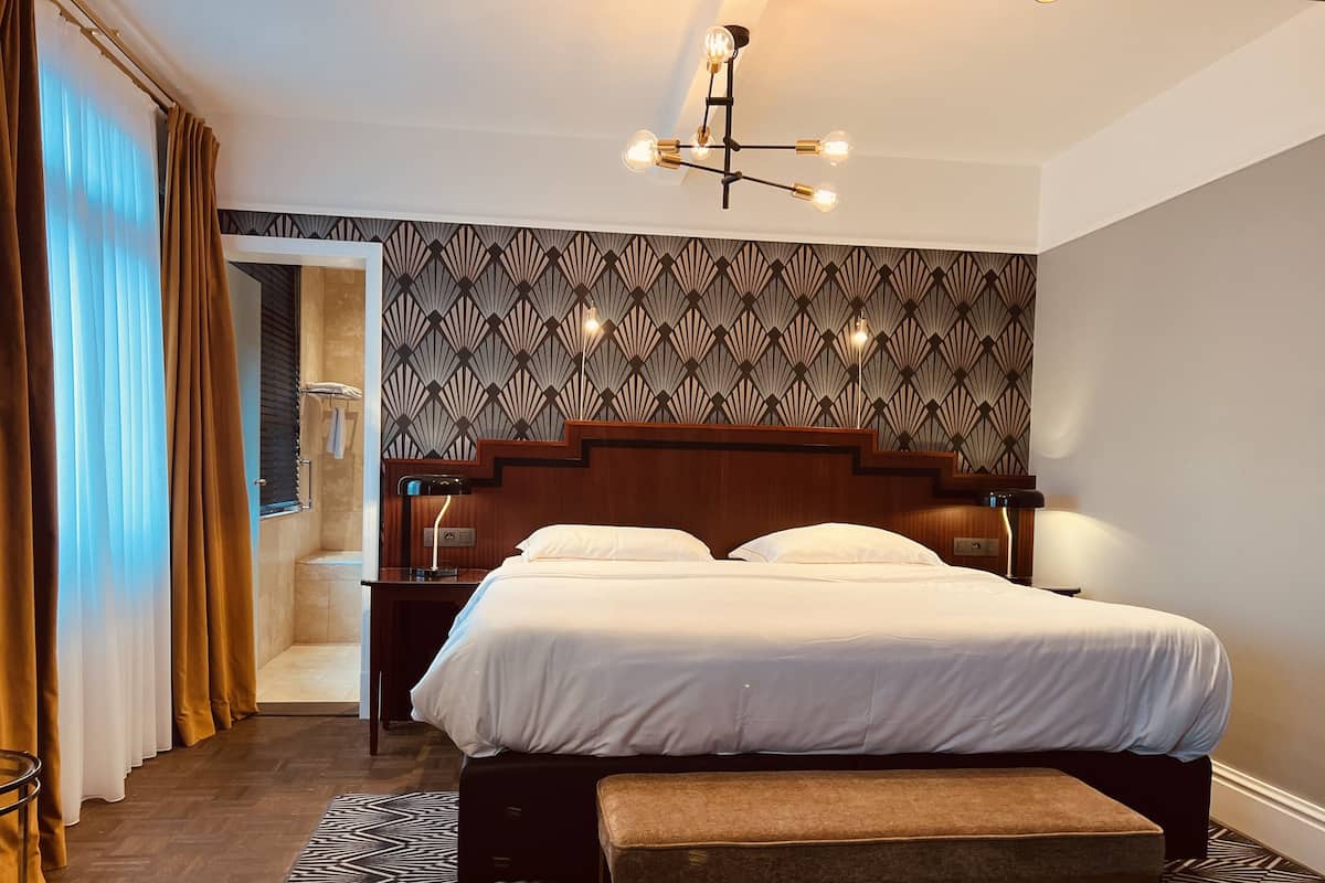 Une chambre d’hôtel avec un grand lit, un banc, une table de chevet avec une lampe, un mur à motifs et un luminaire au plafond.