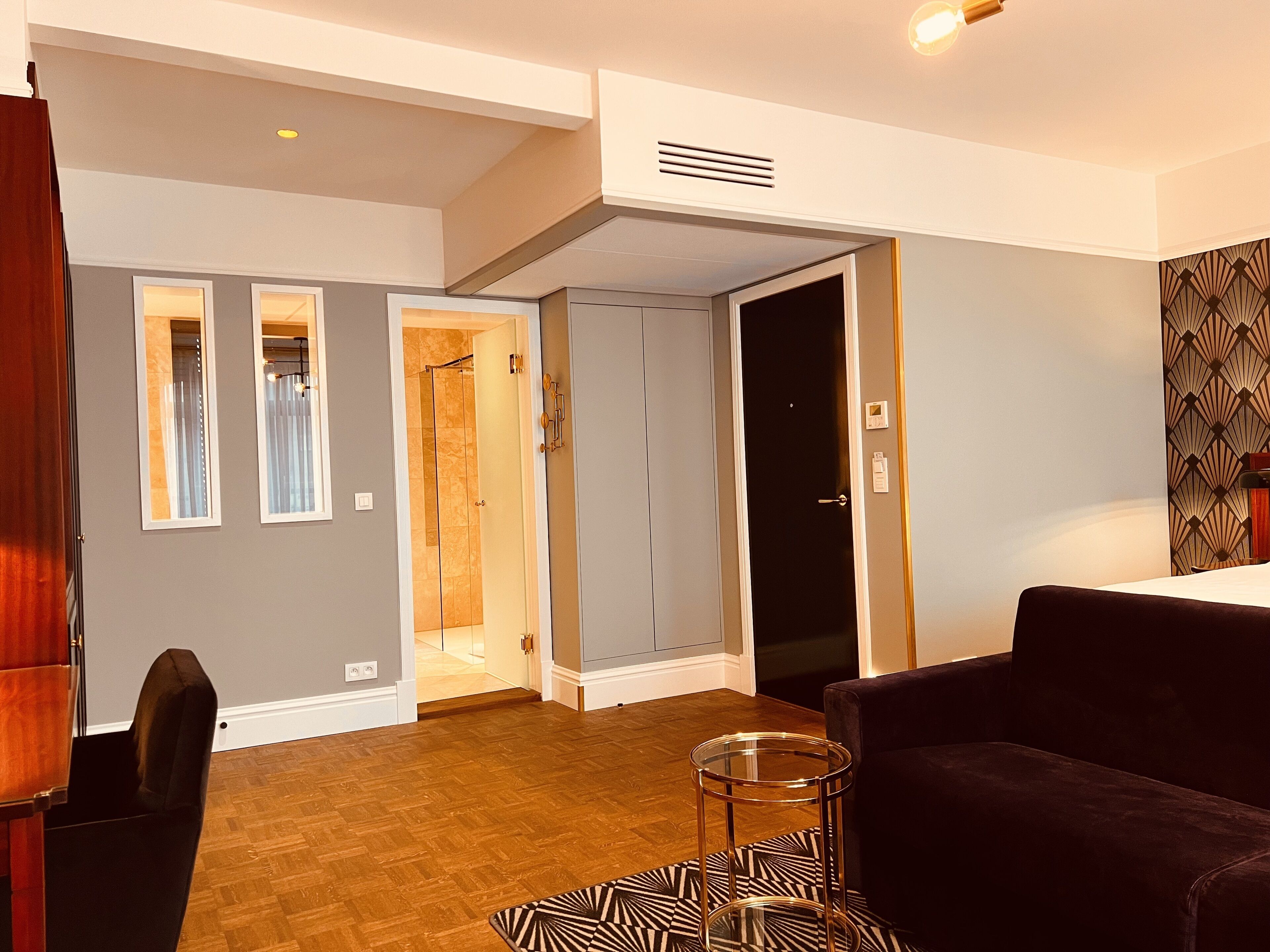 A modern living room with a dark-colored sofa, a glass coffee table, and a patterned rug. There is a door leading to a bathroom with a shower and a mirror. The room has wooden flooring and light gray walls.