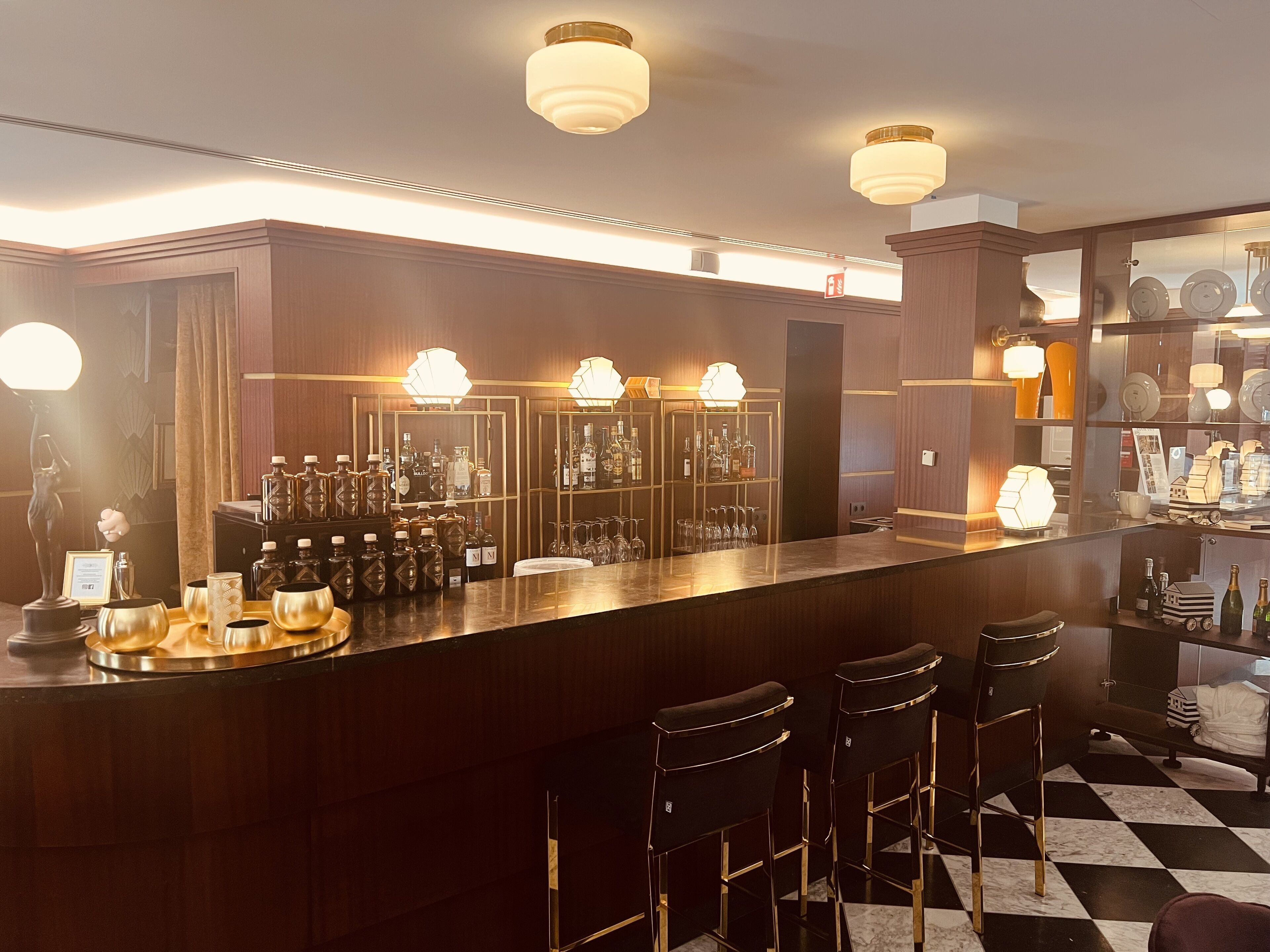Zona de bar con piso a cuadros, taburetes y una barra con botellas y vasos.