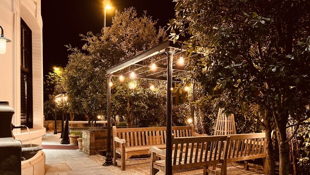 Een buitenruimte met houten banken en een pergola, omringd door bomen en verlicht met lichtslingers.