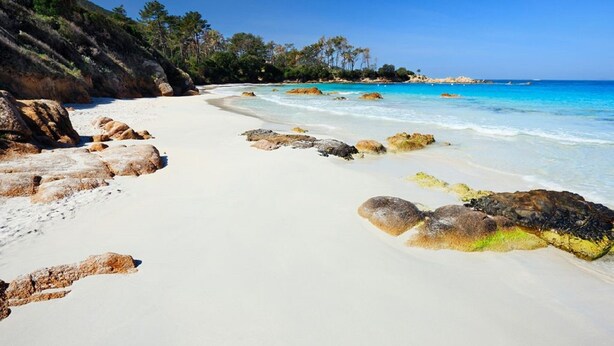 Une plage de sable blanc, avec une eau d’un bleu limpide et quelques rochers éparpillés.
