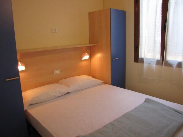 A single bed with a wooden headboard, a blue wardrobe, and a window with curtains.