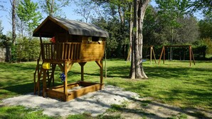 A wooden playhouse with a roof and a ladder, situated on a grassy area with trees and a swing set in the background. - Camping Viareggio (Viareggio)