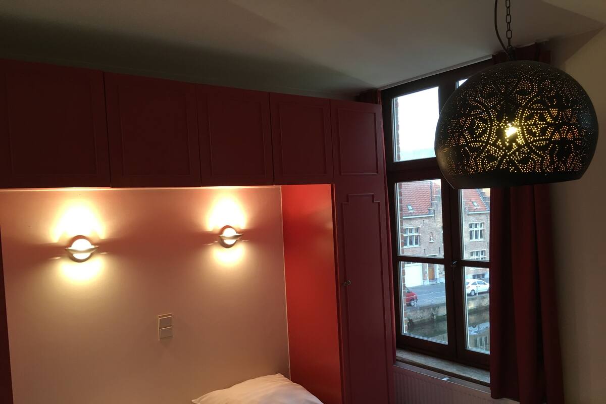 Un dormitorio con una cama, dos luces fijadas en la pared, una ventana con vistas a edificios y una lámpara colgante.