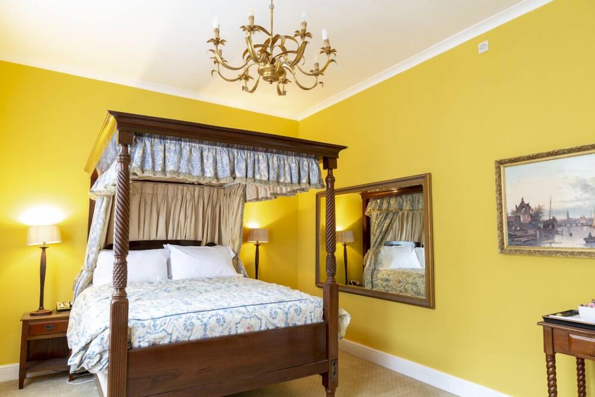 A four-poster bed with a canopy, two bedside lamps, a mirror, and a painting on the wall.