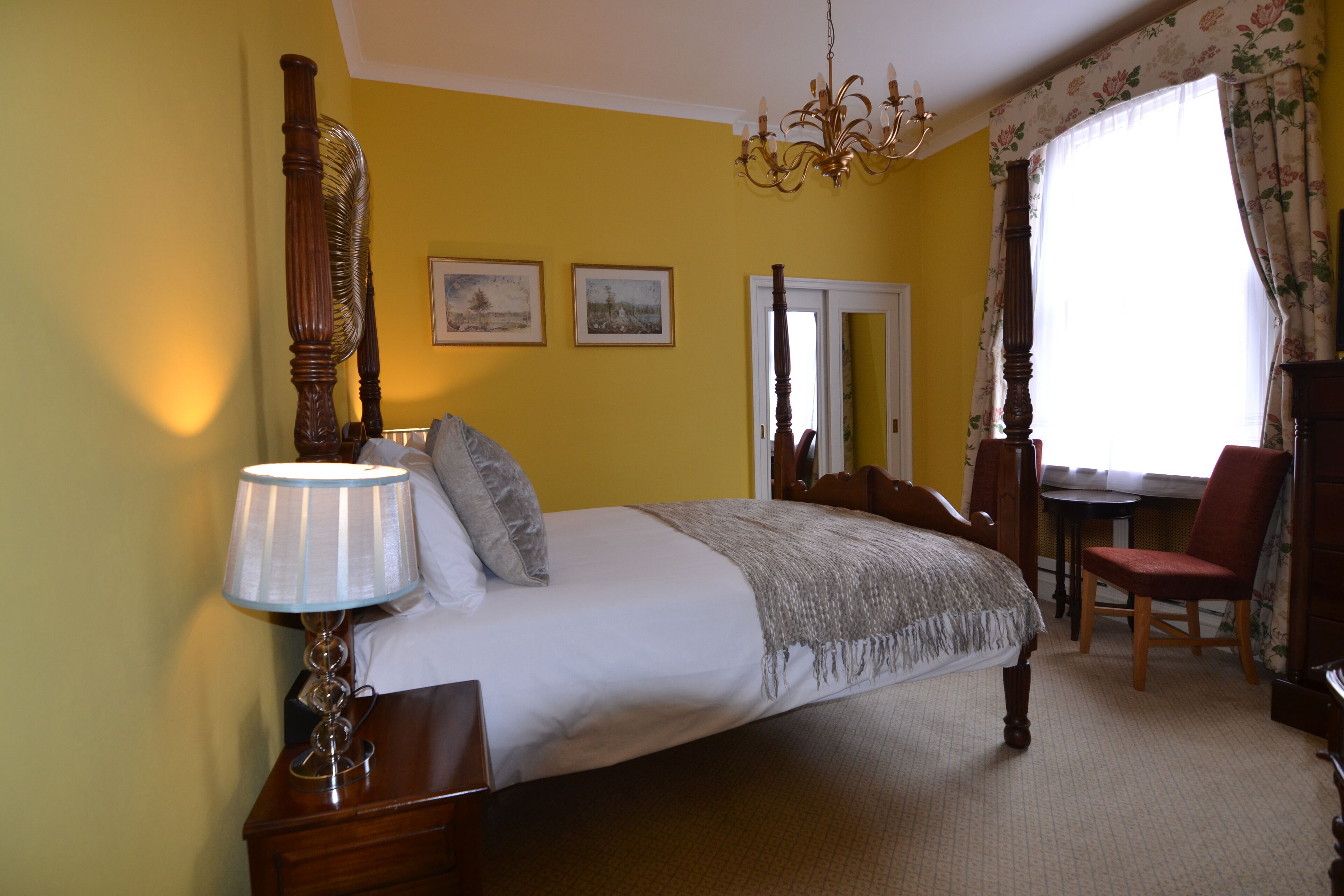 A bedroom with a four-poster bed, a chandelier, a desk with a chair, and two framed pictures on the wall.