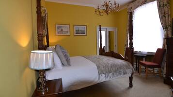 A bedroom with a four-poster bed, a chandelier, a desk with a chair, and two framed pictures on the wall.