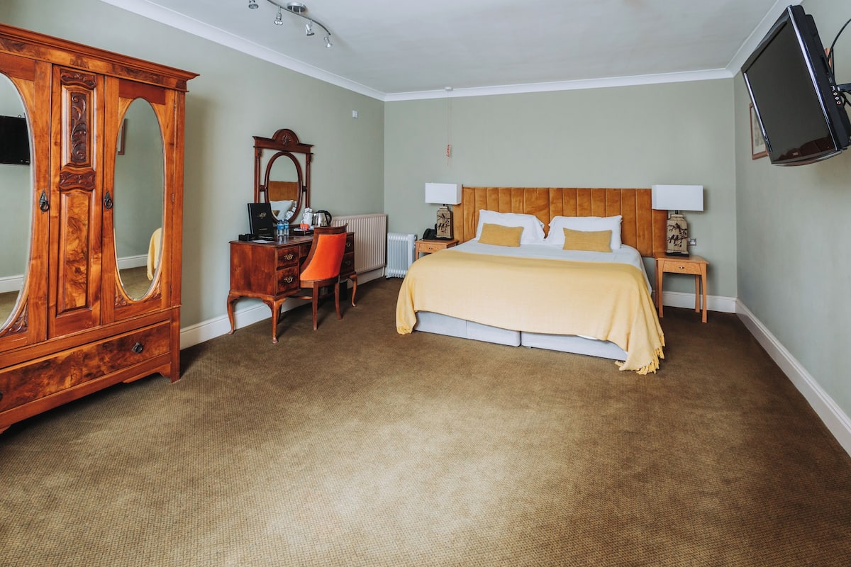 A bedroom with a bed, a wooden wardrobe, a desk with a chair, a television, and a radiator.