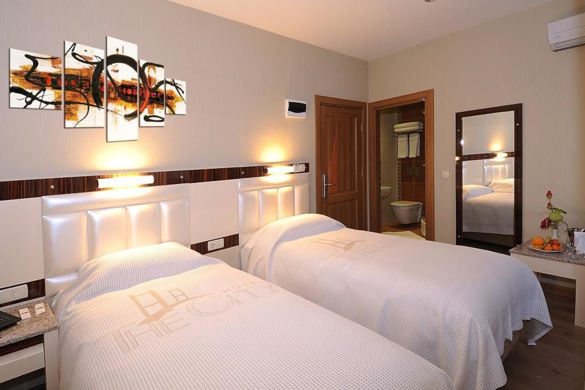 A hotel room with two beds, a large mirror, a wall art piece, and a bathroom visible through an open door.