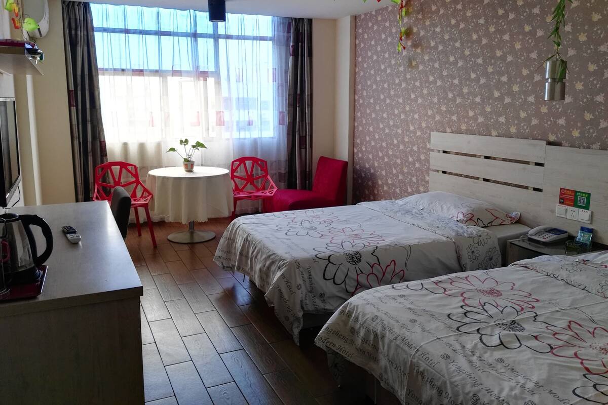 Habitación de hotel con dos camas, un escritorio, una silla, una mesita con una planta y una ventana con cortinas.