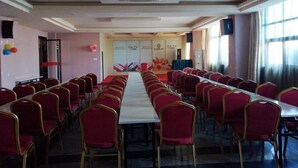 Un salón de conferencias con mesas largas y sillas rojas, una pizarra y un amplio ventanal con cortinas.