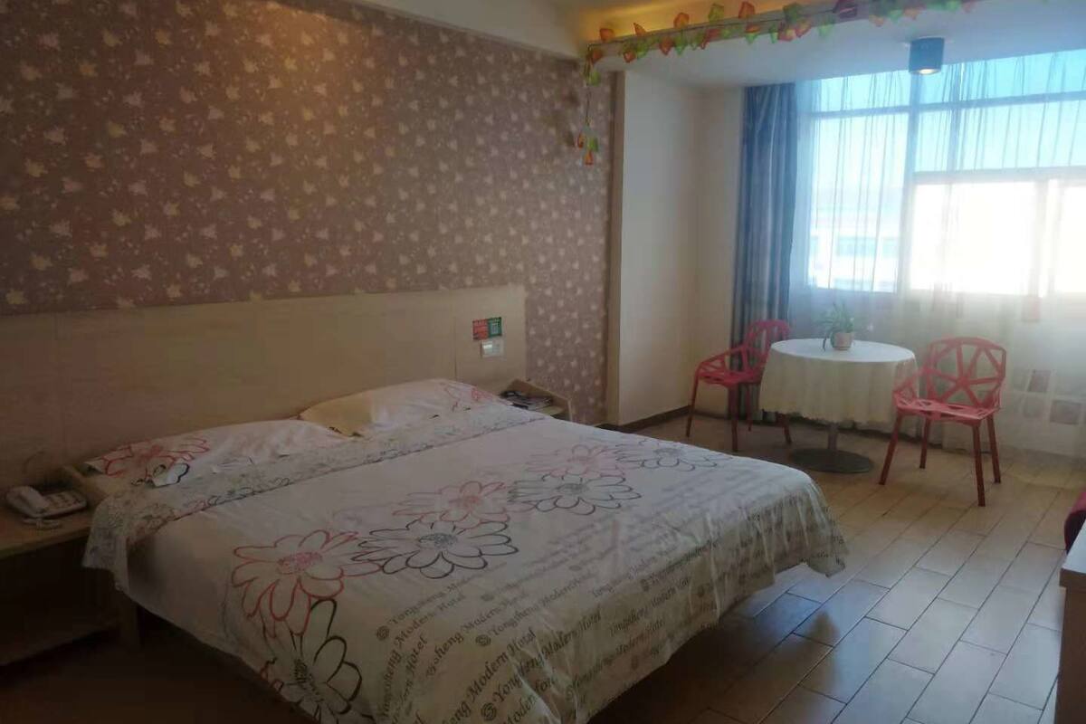Habitación de hotel con cama, una pequeña mesa con sillas, un ventanal con cortinas y una pared con estampado floral.