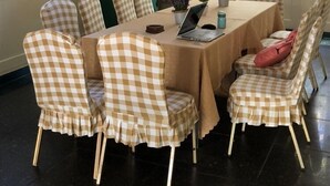 Un comedor con una mesa dispuesta para ocho personas, rodeada de sillas a cuadros. Hay un ordenador portátil sobre la mesa, y un ventanal deja entrar luz natural.