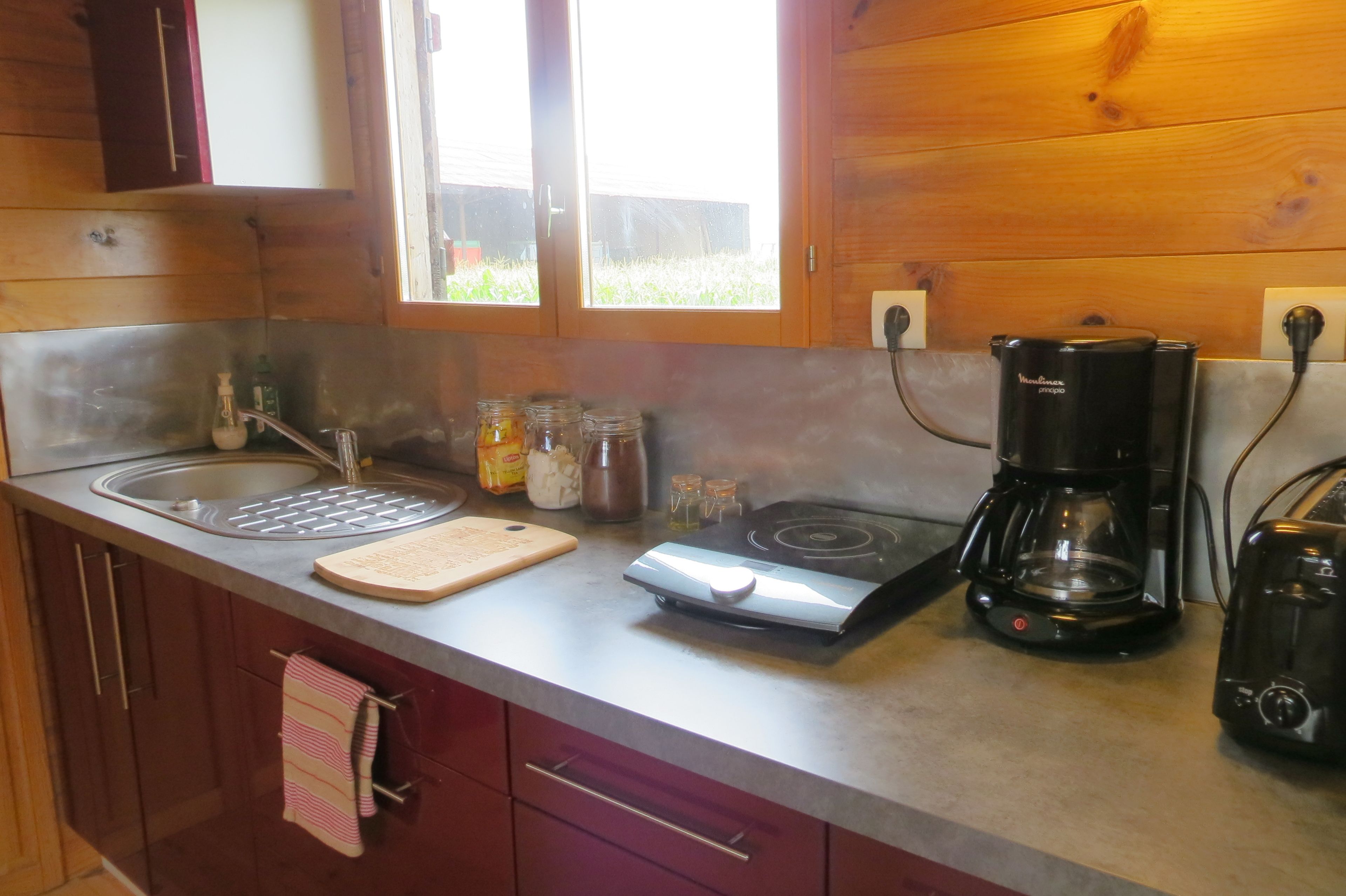 Una cocina con un lavamanos de acero inoxidable, tabla de cortar, estufa, cafetera y frascos sobre la encimera.