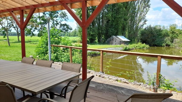 A covered outdoor seating area with a table and chairs, overlooking a pond and a small building. - Roulotte à la Ferme de l'Isle (Avaray)