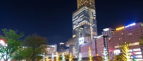 A modern high-rise building with illuminated windows and signage, surrounded by a well-lit urban area with trees and a grassy park.