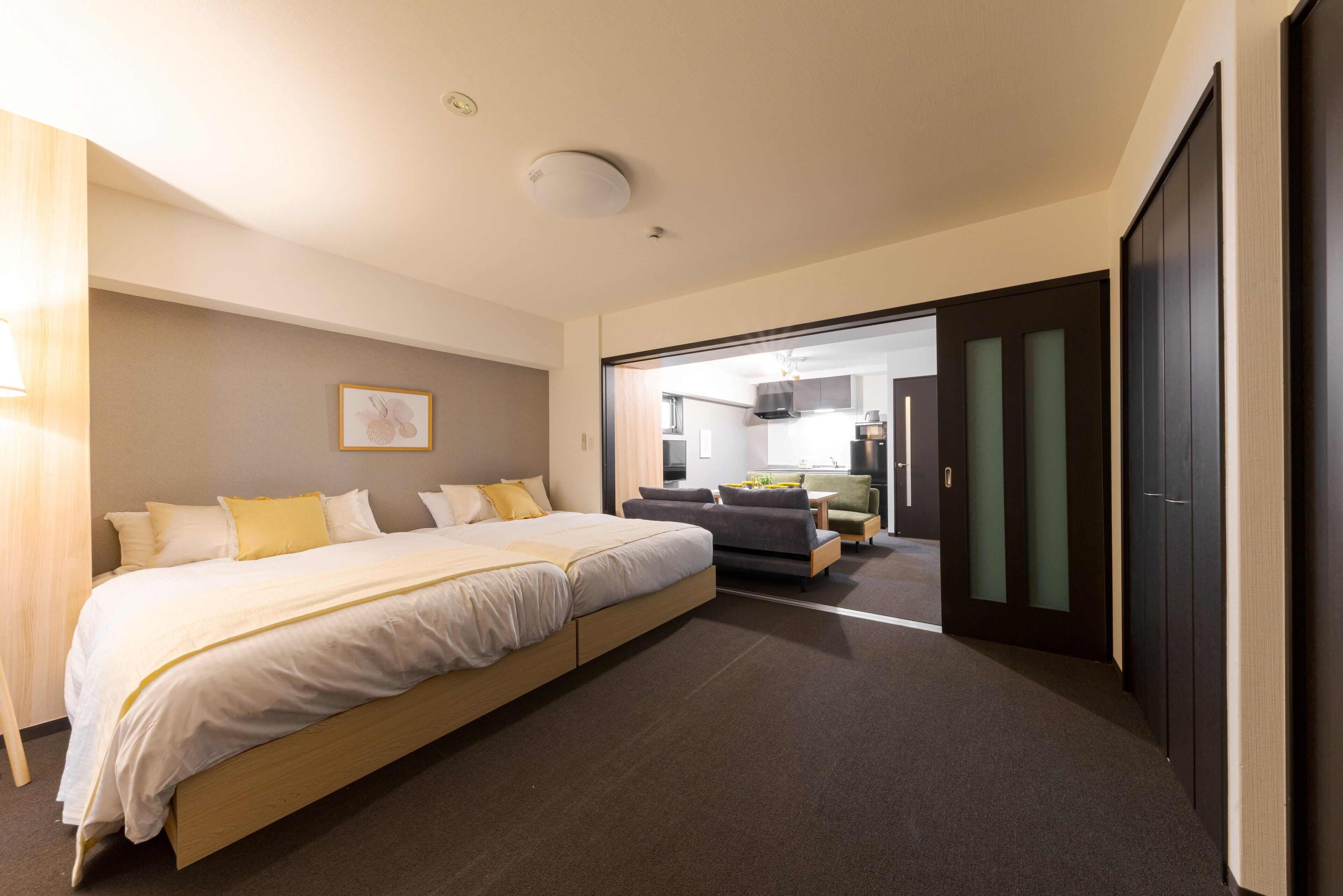 Une chambre d’hôtel moderne avec un grand lit, un canapé et un bureau. Une porte donne accès à un coin cuisine.