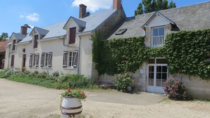 A row of traditional stone houses with pitched roofs, a gravel driveway, and a barrel planter with flowers in the foreground. - Chambres d'hotes Domaine de l'Isle (Avaray)