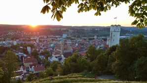 A panoramic view of a town with a prominent tower and a church, surrounded by greenery and a pathway with people walking.