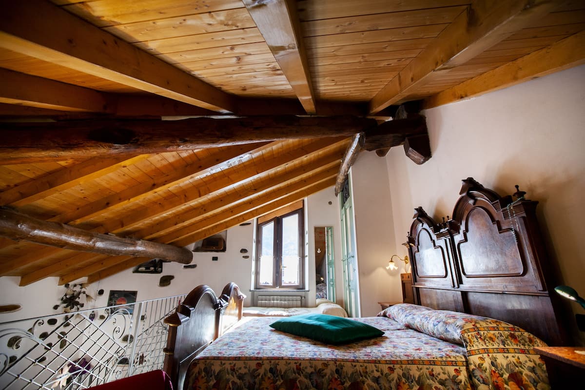 Une chambre avec un plafond en bois incliné, un lit avec une literie à motifs floraux, une tête de lit en bois et une fenêtre laissant entrer la lumière naturelle.