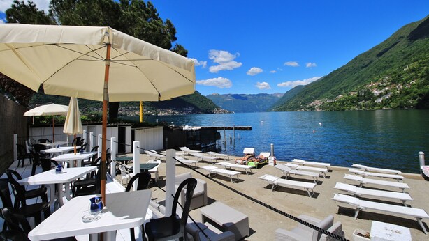 A terrace with white tables and chairs, a large umbrella, and a view of a lake with mountains in the background. - Antica Molina (Faggeto Lario)