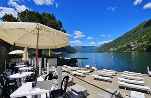 Une terrasse avec des tables et des chaises blanches, un grand parasol et une vue sur un lac, avec des montagnes en arrière-plan.