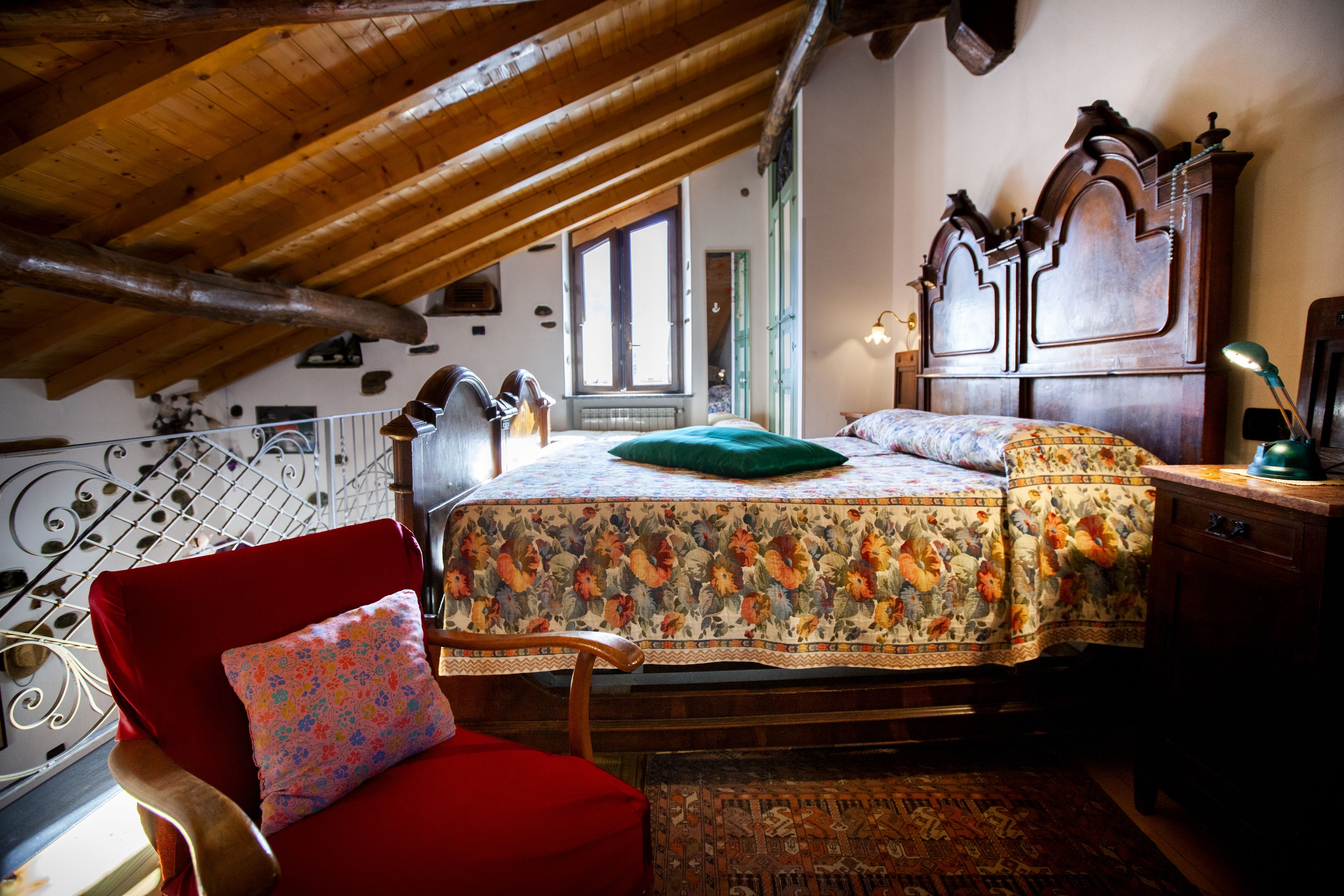 Ein Schlafzimmer mit Holzbalkendecke, einem blumigen Bettbezug, einem roten Sessel und einem Nachttisch aus Holz.