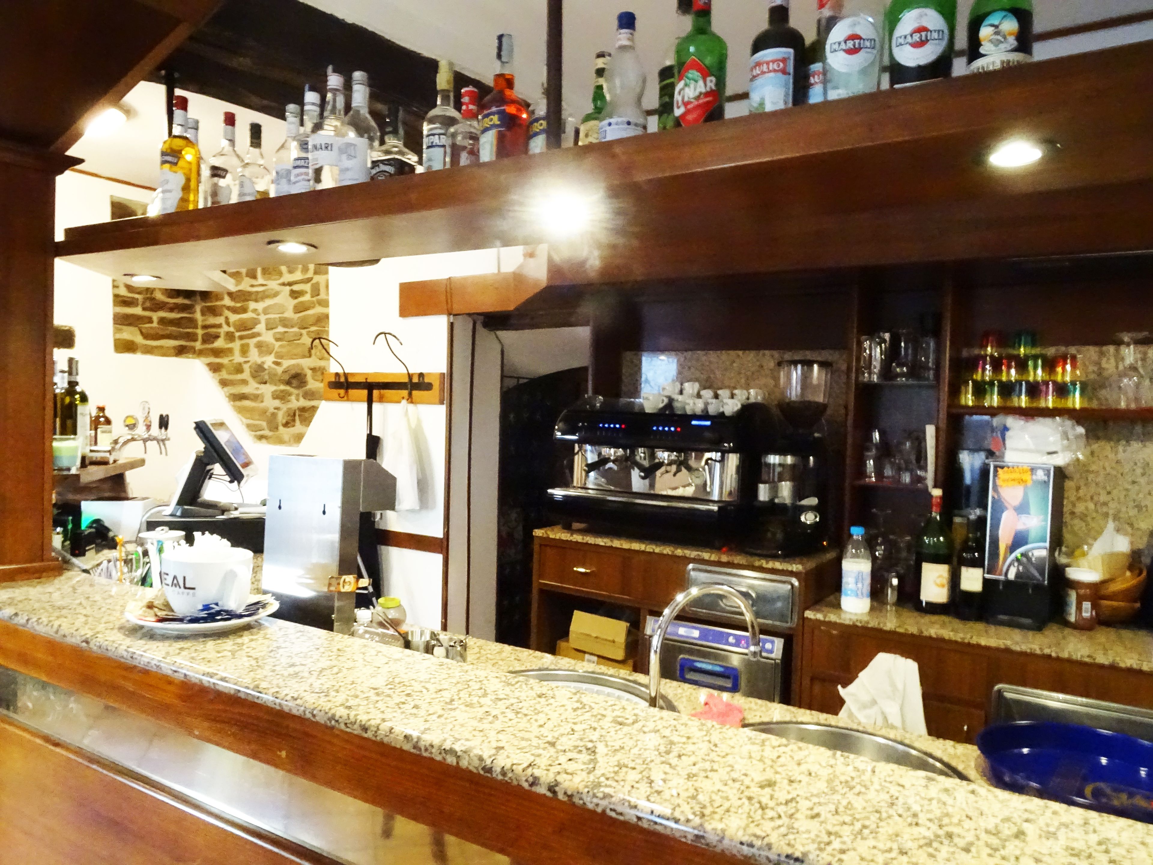 Uma área de bar com máquina de café, prateleiras de garrafas de bebidas e uma parede de pedra ao fundo.