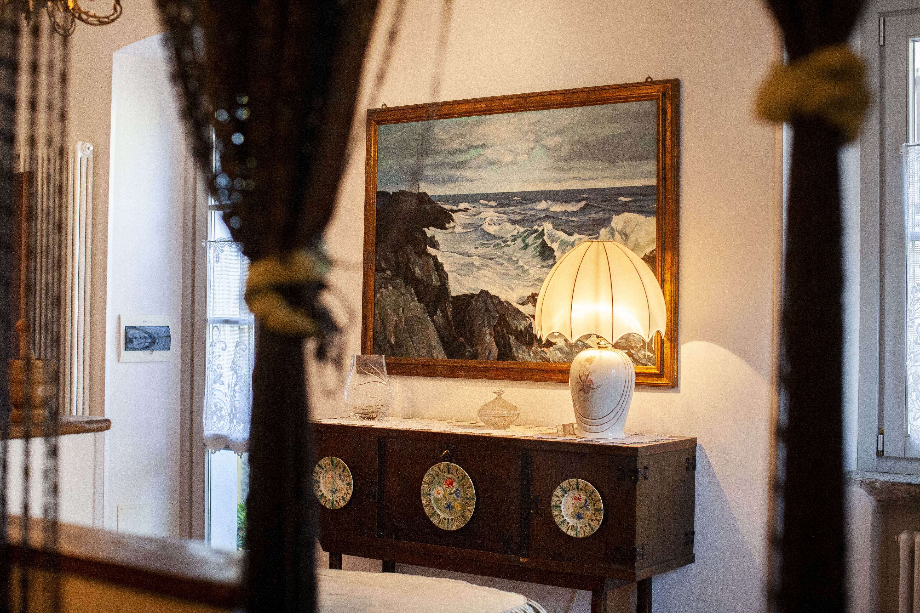 Une chambre à coucher avec un lit, une table de chevet avec une lampe, un tableau représentant un paysage marin et une fenêtre avec des rideaux.