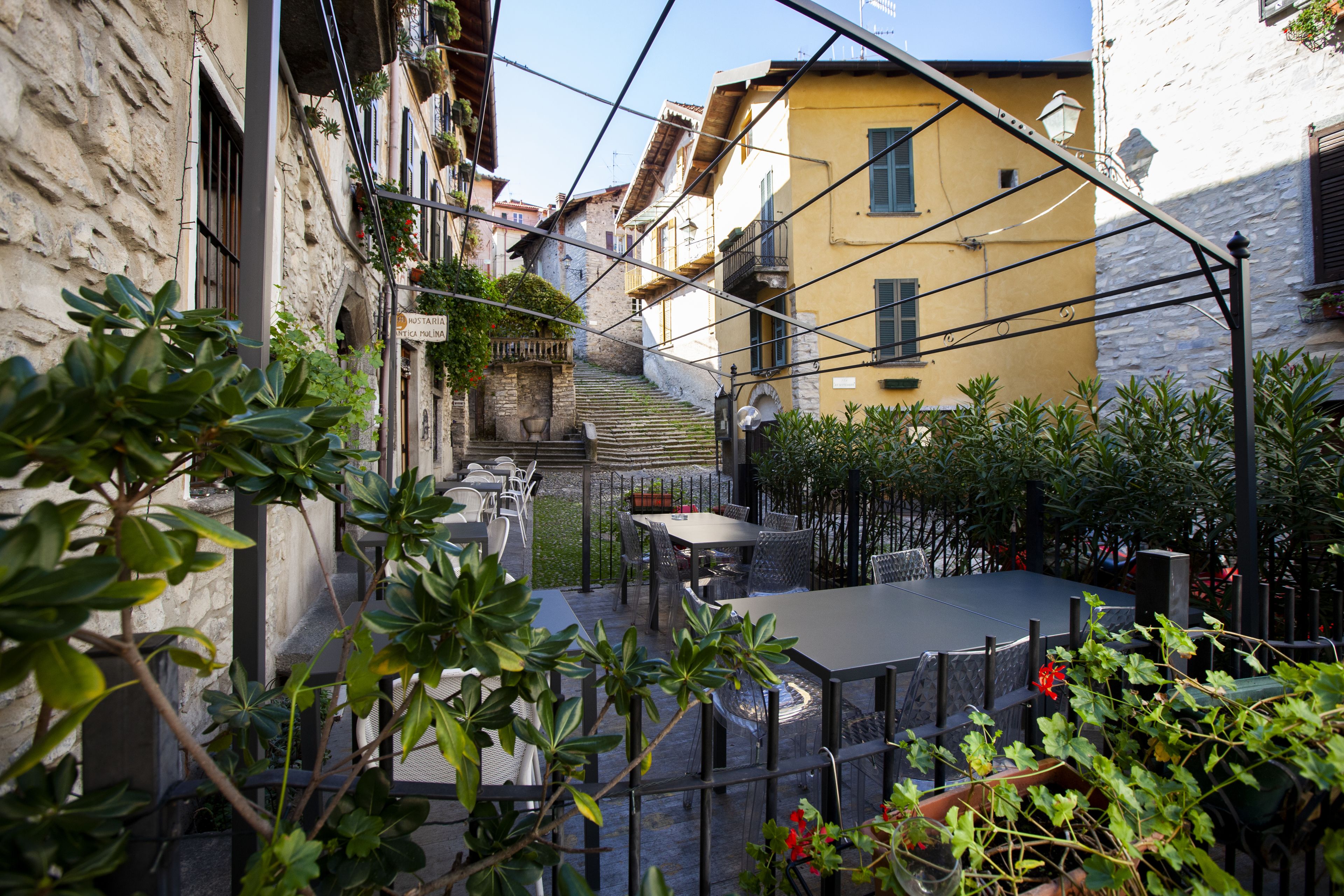Una calle estrecha flanqueada por edificios, con mesas al aire libre y una escalera que sube a un nivel superior.