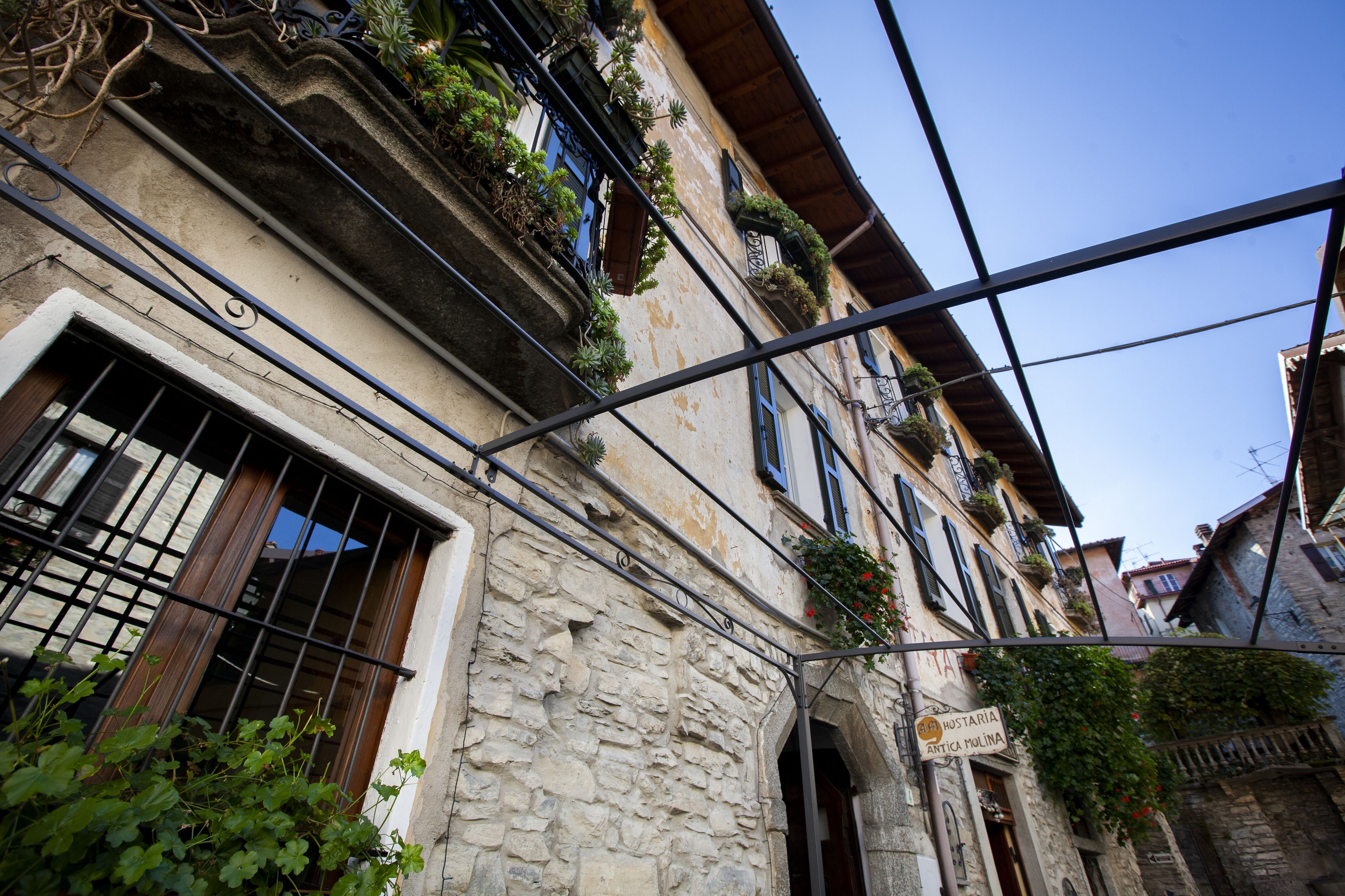 Una stretta via con edifici in pietra, balconi fioriti e una insegna che indica un ristorante.