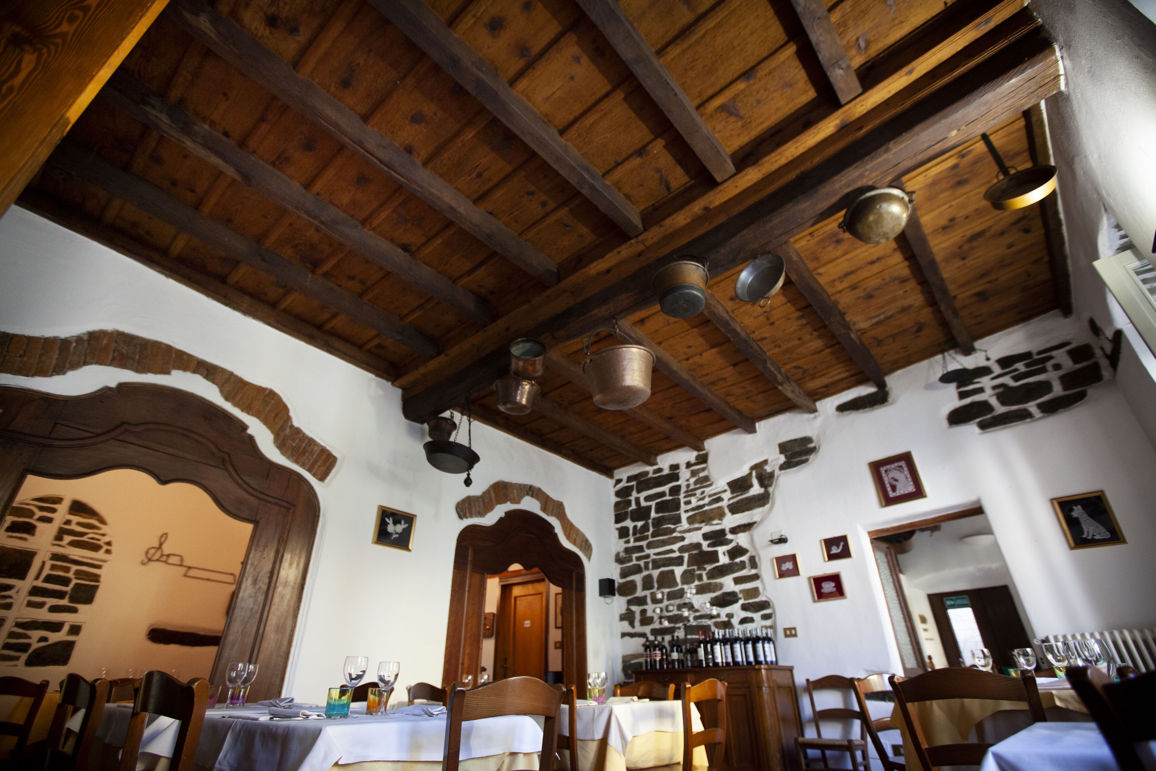 Un coin repas rustique avec des poutres en bois et des murs en pierre, des tables dressées avec des verres à vin et des assiettes, et un mur en pierre orné de tableaux.
