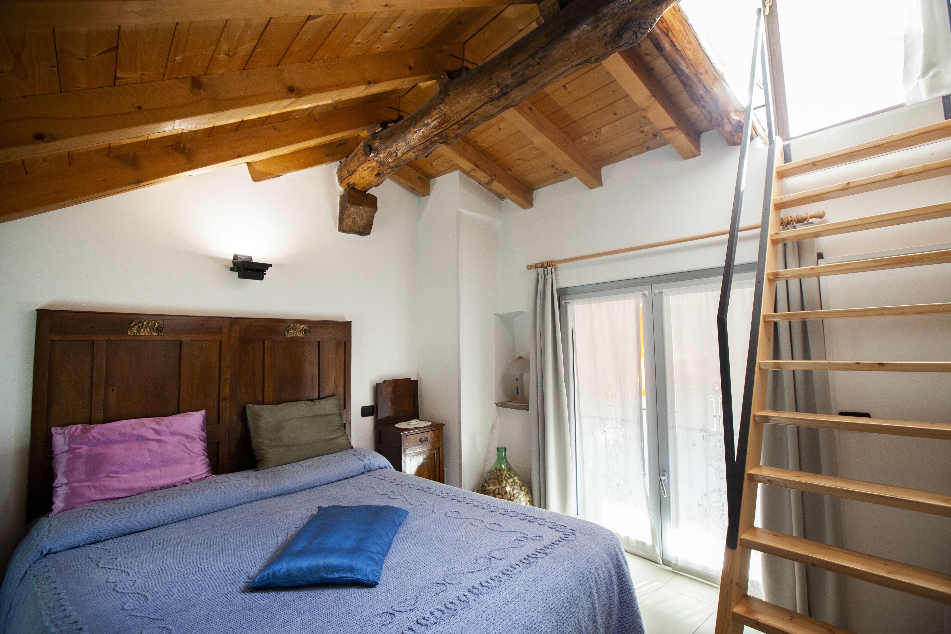 A bedroom with a wooden ceiling, a bed with a blue pillow, a wooden ladder, and a window with curtains.