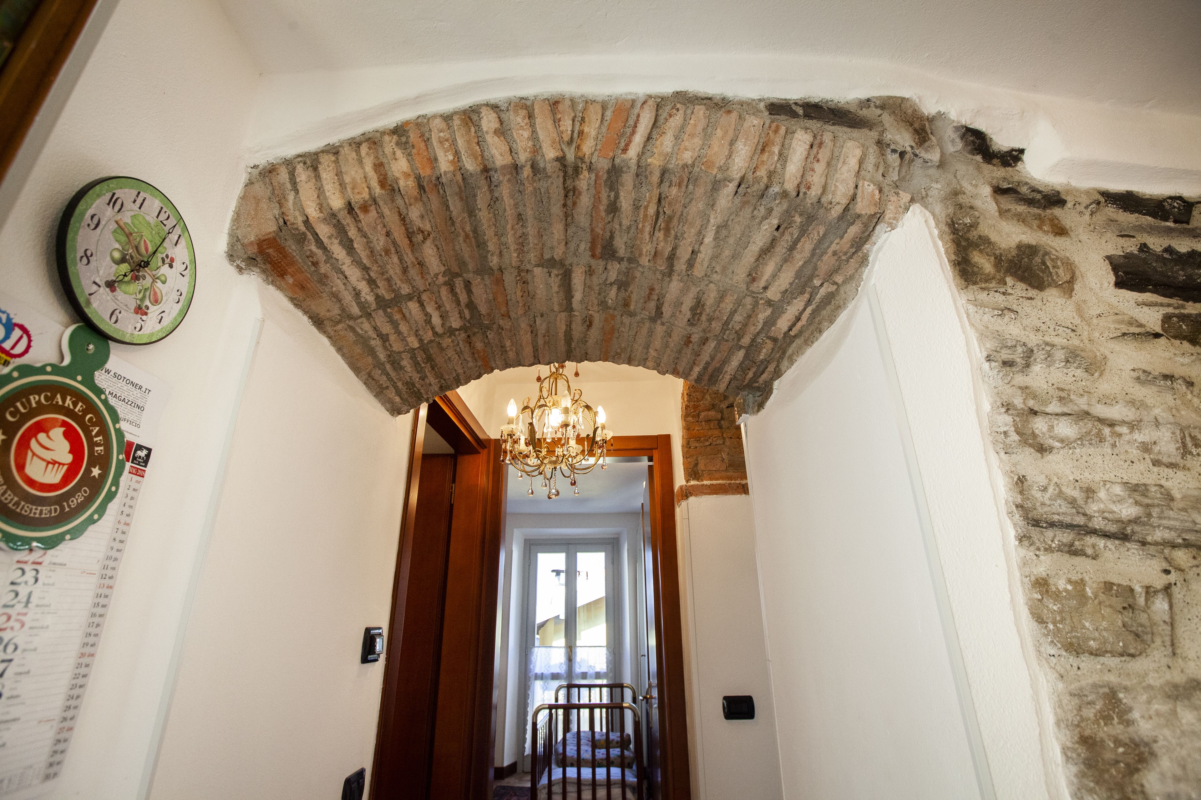 A hallway with a brick archway, a chandelier, and a clock on the wall.