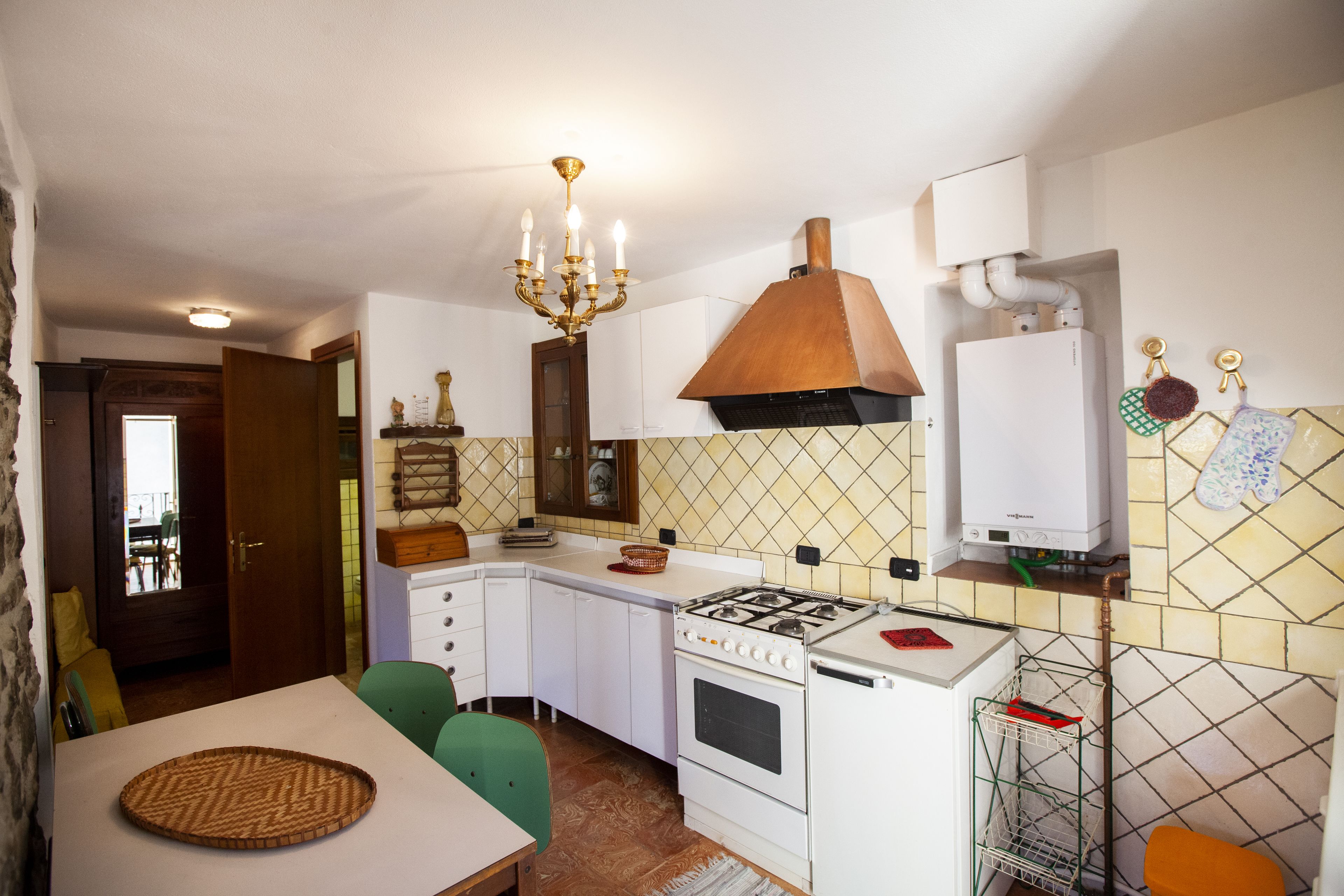 Una cocina compacta con gabinetes blancos, estufa a gas, horno empotrado, un colgador para ollas y una zona de comedor con mesa y sillas.