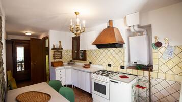 Una cocina compacta con gabinetes blancos, estufa a gas, horno empotrado, un colgador para ollas y una zona de comedor con mesa y sillas.